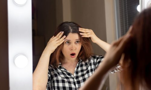 Horrified young woman looking in the bathroom mirror staring open mouthed at the first grey hair on her scalp, a first sign of ageing, or noticing that she is suffering from dandruff
